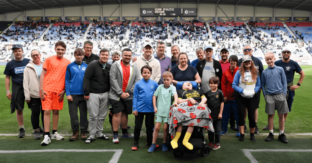 A group of people, including children and adults, pose for a photo on a sports field with a stadium filled with spectators in the background. One child is in a wheelchair.