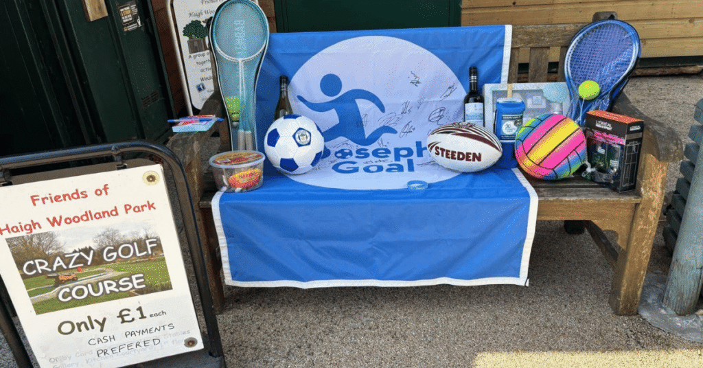 Fundraising stall for Joseph’s Goal with a signed blue banner, footballs, rugby balls, tennis rackets and other sports items, alongside a £1 crazy golf course sign.