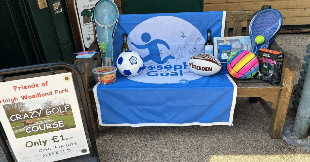 Fundraising stall for Joseph’s Goal with a signed blue banner, footballs, rugby balls, tennis rackets and other sports items, alongside a £1 crazy golf course sign.